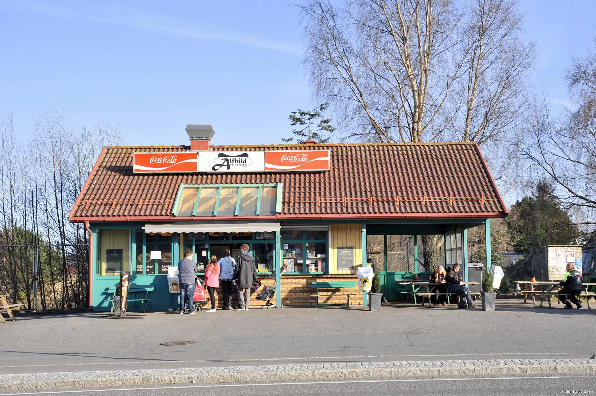 Den gule kiosken med grønne lister og rødt teglstak i Nansetgata. Skiltet «Alfhild på Hovland» over luka, kunder ved disken og folk ved bordene under taket.